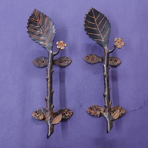 May include: A pair of dark metal door handles shaped like tree branches with textured leaves and small flowers. Each handle features two horizontal mounting plates with pre-drilled screw holes, finished in a dark brown tone with copper highlights.