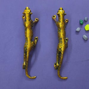 Puede incluir: Dos manijas de puerta de metal color dorado con forma de leopardo, con manchas detalladas y cuerpos alargados. Cada manija tiene orificios de montaje en la parte superior e inferior. A la derecha hay cuatro piedras decorativas pequeñas y lisas.