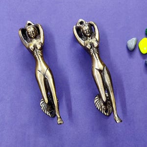 Może przedstawiać: Dwie identyczne mosiężne klamki do drzwi w kształcie stylizowanych postaci z rękami uniesionymi nad głową. Każda figurka ma gładkie, wydłużone ciało i podstawę o fakturze wachlarza. Obok znajdują się cztery małe ozdobne kamienie.