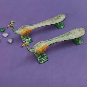 Peut inclure: Deux poignées de porte en métal en forme de paon avec des finitions peintes en vert et doré. Chaque poignée présente une longue queue texturée s'étendant du corps de l'oiseau, avec des plaques de fixation à la base. Quatre petites pierres lisses.
