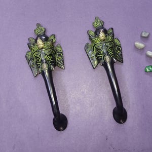 Puede incluir: Un par de manijas de puerta de metal negro con un diseño ornamentado de cabeza de elefante con detalles en verde y dorado. Cada manija tiene un agarre largo y curvo en forma de trompa y una sección superior decorativa con piedras.