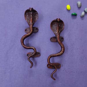 May include: Two bronze-coloured metal door handles shaped like cobras with raised hood details and intricate engraved patterns. Each handle features two oval mounting plates with screw holes, positioned vertically to form a serpentine silhouette.