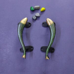May include: A pair of metal door handles shaped like fish with green scale patterns and gold-toned heads and tails. Each handle features two black mounting plates with screw holes. Four small, smooth, multi-colored stones are placed above them.
