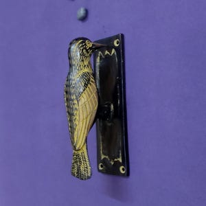 May include: A black and yellow bird-shaped door knocker mounted on a rectangular black metal plate. The bird figure features detailed etched lines and a pointed beak, with the metal plate containing four circular screw holes at the corners.