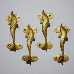 May include: A set of four gold-colored metal cabinet handles, each shaped like a stylized fish with detailed scales and fins. Each handle features two oval mounting plates with pre-drilled screw holes at the top and bottom for secure attachment.