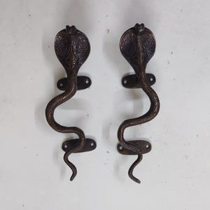 May include: A pair of dark bronze-toned metal door handles shaped like upright cobras. Each handle features an intricate, embossed pattern along the snake's body and hood, with two oval mounting plates containing screw holes at the top and bottom.