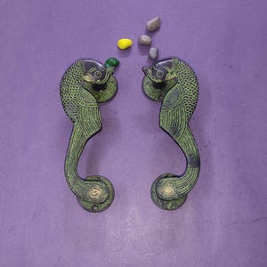 May include: A pair of dark metal door handles featuring an intricate peacock design with engraved feather patterns and a green finish. The handles are curved and symmetrical, with small decorative stones placed between them on the surface.