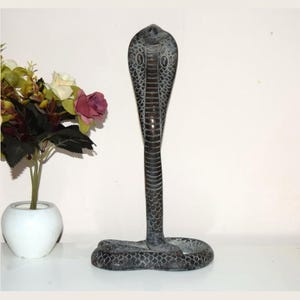May include: A dark gray, upright cobra snake figurine featuring a textured scale pattern across its body and hood. The sculpture is crafted with a flared hood and a coiled base, displaying detailed carvings that mimic the natural appearance of a cobra.