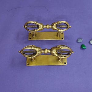 Puede incluir: Dos manijas de puerta de latón a juego, cada una con un diseño central inspirado en el vajra con intrincado trabajo en metal. Las manijas están montadas sobre placas rectangulares de latón con orificios para tornillos preperforados.