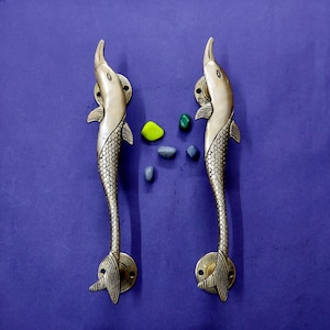 May include: A pair of vertical brass door handles shaped like stylized fish. Each handle features a smooth head, a textured scale pattern along the body, and a flared tail fin. The handles are mounted on circular plates with visible screw holes.