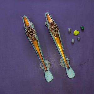 May include: Two elongated door handles featuring a central brass-toned lion head relief. The handles have a distressed light blue finish on the edges and circular mounting plates at each end, showcasing a weathered and textured appearance.