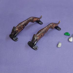 May include: Two dark bronze-toned metal door handles shaped like prowling tigers. Each handle features a detailed, elongated feline body with a textured, striped surface and two mounting plates with screw holes at the head and tail ends.