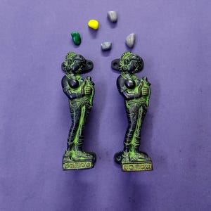 May include: Two identical dark metal door handles featuring a standing figure with green highlights, each with mounting plates at the top and bottom. Above the handles are five small, smooth, multi-colored stones in yellow, green, and gray.
