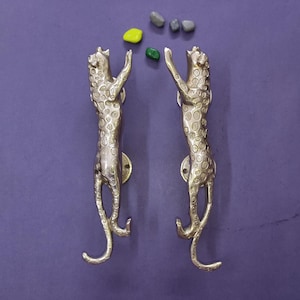 Peut inclure: Une paire de poignées de porte en métal doré en forme de léopards bondissants, ornées de taches circulaires texturées sur tout le corps. Chaque poignée est orientée verticalement avec une queue courbée et une plaque de fixation circulaire.