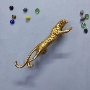 Op de afbeelding: Een goudkleurige metalen handgreep of deurknop in de vorm van een luipaard met een gestructureerd vlekkenpatroon over het lichaam. De luipaard is afgebeeld in een springende houding met de voorpoten naar voren gestrekt. Diverse kleine glazen steentjes liggen verspreid.