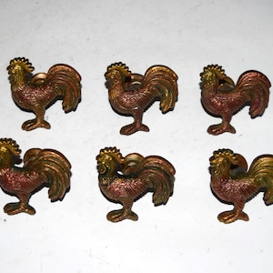 Può includere: Un set di sei pomelli metallici per armadietti a forma di gallo. Ogni pomello presenta una finitura testurizzata e iridescente con toni rame e bronzo, raffigurante un gallo in piedi con cresta prominente, piumaggio dettagliato e coda curva.