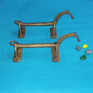 May include: A pair of metallic brass door handles shaped like stylized horses. Each handle features a long, textured body with a curved neck and head, supported by two rectangular mounting plates with pre-drilled holes for installation.