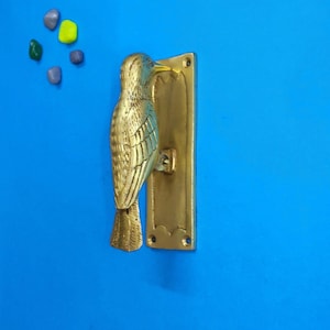 May include: A brass door knocker shaped like a bird, featuring a rectangular mounting plate with screw holes at the top and bottom. The bird is depicted in a vertical position with detailed wing textures and a pointed beak, attached to the plate.