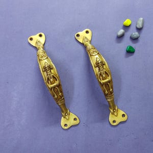 May include: A pair of gold-coloured metal door handles featuring an embossed figure in the centre of each handle. Each handle has a curved body with decorative patterns and two flat mounting plates at the ends, each with two circular screw holes.