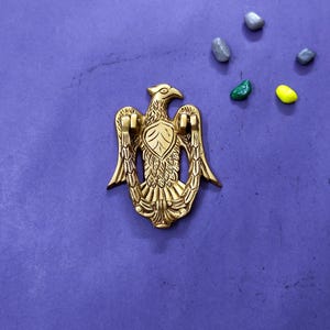 May include: A gold-coloured metal door knocker shaped like an eagle with spread wings. The eagle features detailed feather engravings and a central shield design on its chest. The wings curve downwards to form the handle of the knocker, which is positioned against a solid purple background.