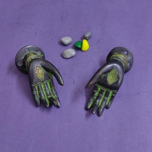 May include: Two dark metal wall hooks shaped like open hands with green patina accents on the fingers and palms. Each hook features a circular mounting base with screw holes and decorative etched patterns on the wrist and palm areas.