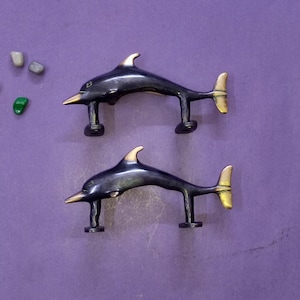 May include: Two metal dolphin-shaped cabinet pulls or handles. Each piece features a dark, polished finish on the body with contrasting metallic gold-toned accents on the nose, dorsal fin, and tail. Both dolphins have two mounting posts underneath.