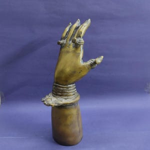 May include: A bronze-colored metal sculpture of an upright hand with fingers slightly spread. The wrist is adorned with multiple stacked bracelets, and each finger features a ring. The sculpture has a smooth, cylindrical base and a metallic finish.