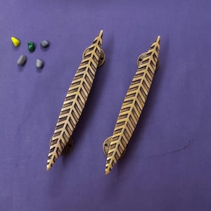 May include: Two elongated brass door handles featuring a detailed, symmetrical leaf-like or feather-like pattern etched into the surface. Each handle is secured by two circular mounting plates with visible screw holes, presented alongside five small stones.