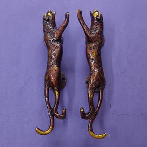 May include: A pair of vertical door handles shaped like standing leopards. The metal handles feature a dark bronze finish with polished gold-toned accents on the leopard spots, faces, and tails. Each handle has a circular mounting plate at the back.