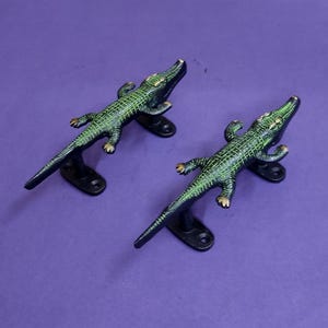 May include: Two metal door handles shaped like crocodiles. Each handle features a textured green and black finish with gold-toned accents on the eyes and claws. The handles are mounted on black oval plates with two screw holes for attachment.
