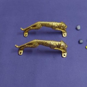 May include: Two gold-colored metal cabinet handles shaped like tigers in a pouncing position. Each handle features engraved tiger stripes and two mounting points with pre-drilled holes at the front and back paws for secure attachment to furniture.