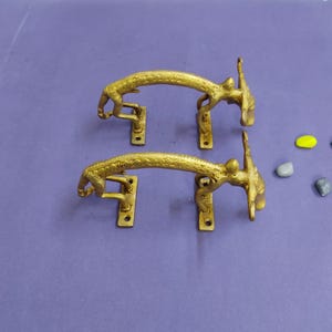May include: Two gold-colored metal door handles shaped like stylized animals. Each handle features a textured, arched body with four small rectangular mounting plates, each containing a single screw hole for attachment to a door or cabinet surface.