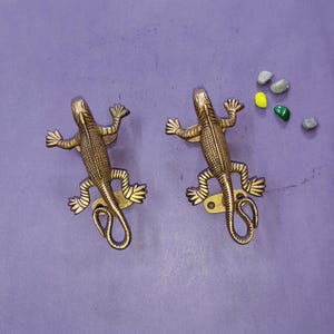 May include: Two identical brass lizard-shaped wall hooks with textured scales and curled tails. Each lizard features a small metal mounting plate with two screw holes near the hind legs, finished in a metallic gold tone with dark antique detailing.