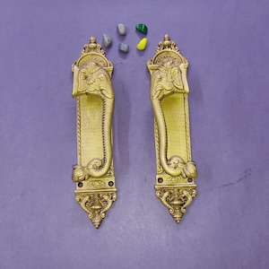 May include: A pair of vertical brass door handles featuring an embossed elephant head design at the top and a curved trunk forming the grip. The rectangular backplates have a dotted texture and ornate decorative detailing at the top and bottom ends.