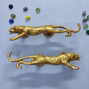 Peut inclure: Deux poignées de porte en métal doré en forme de léopards bondissants avec des taches détaillées et la gueule ouverte. Les poignées sont placées horizontalement, l'une au-dessus de l'autre, et entourées de plusieurs petits galets en verre colorés.