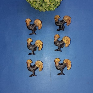 Può includere: Un set di sei pomelli per armadietti a forma di gallo disposti in due colonne. Ogni pomello presenta un corpo scuro e testurizzato con una finitura oro metallizzato a contrasto su testa, collo e piume della coda.