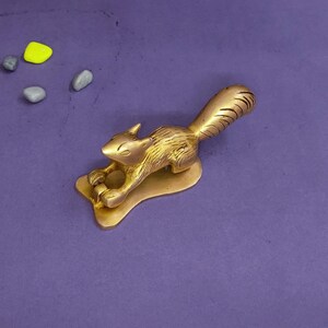 Op de afbeelding: Een metallic goudkleurig koperen eekhoornfiguur in een gehurkte houding, met gedetailleerde vachttexturen en een gebogen staart. De eekhoorn is bevestigd op een platte, onregelmatig gevormde basis. Vier kleine, gladde steentjes in groen, geel en grijs liggen in de buurt.