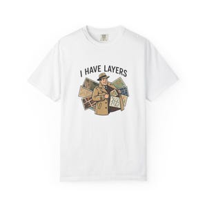 May include: A white t-shirt featuring a graphic of a person in a tan trench coat and fedora hat opening the coat to reveal various maps. The text I HAVE LAYERS is printed in black above the illustration, which is centered on the chest area.