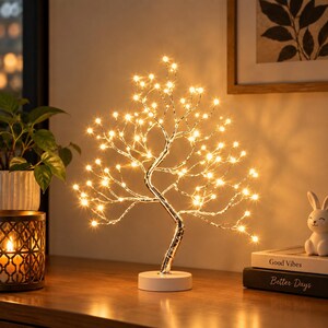 Op de afbeelding: Een decoratieve boomlamp met een zilverkleurige stam en takken versierd met talloze kleine, warmwitte LED-lampjes. De boom is gemonteerd op een ronde witte voet en verspreidt een zacht, sfeervol licht door de gehele structuur.
