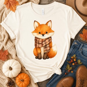 Könnte beinhalten: Ein cremefarbenes Kurzarm-T-Shirt mit einer zentralen Grafikillustration eines kleinen orangefarbenen Fuchses, der einen braun-beigen Schal mit Karomuster trägt. Der Fuchs hat spitze Ohren, dunkle Augen und einen buschigen Schwanz.