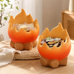 Könnte beinhalten: Zwei Keramikschalen in Form stilisierter Lagerfeuer mit orange-gelben, flammenartigen Rändern und braunen, holzartig texturierten Sockeln. Jede Schale enthält kleine, verpackte Süßigkeiten und steht auf einer hellen Holzoberfläche.