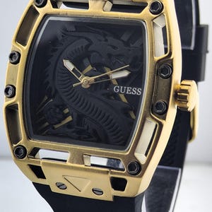 May include: A wristwatch with a black silicone strap and a gold-tone metal case featuring an open-frame design with black screws. The black dial displays a raised dragon motif, gold-tone hands with white accents, and the word GUESS in white.