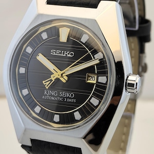 May include: A King Seiko automatic wristwatch with a black dial featuring horizontal lines, gold-tone hands, and white hour markers. The watch has a silver-tone stainless steel case and a black textured leather strap with a date window at the 3 o'clock position.