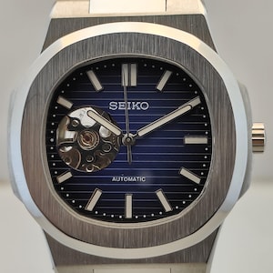 May include: A silver-toned stainless steel wristwatch with a dark blue horizontal striped dial. The watch features a circular open-heart aperture at the nine o'clock position, silver baton indices, and the text SEIKO and AUTOMATIC on the face.
