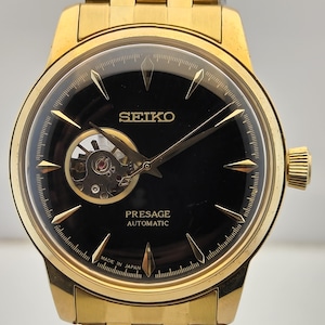 May include: A gold-tone Seiko Presage automatic watch with a black dial. The watch features a round case, a gold-tone metal link bracelet, and an open-heart aperture at the nine o'clock position. The dial displays the text SEIKO, PRESAGE, AUTOMATIC, and MADE IN JAPAN.