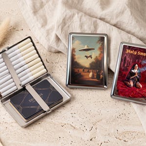 May include: Three rectangular metal cigarette cases with silver frames. One case is open, holding white cigarettes and dark blue business cards. The other two cases are closed, featuring printed images of a UFO with a cow and a nun with the text Holy Smokes.