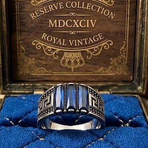 May include: A silver ring featuring three rectangular blue stones set in a row, flanked by engraved geometric patterns on the band. The ring is displayed inside a wooden box with a blue velvet interior and the text Reserve Collection MDCXCIV Royal Vintage.