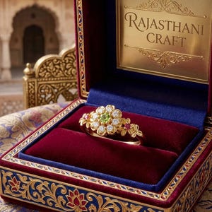 Könnte beinhalten: Ein goldener Ring mit einem zentralen grünen Edelstein, umgeben von weißen Steinen in Blumenform und kleinen roten Akzentsteinen, präsentiert in einer mit Samt ausgekleideten Schatulle mit dem Schriftzug RAJASTHANI CRAFT im Deckel.