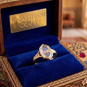 May include: A gold ring with a hexagonal setting featuring a clear center stone and a horizontal band of small red and yellow gems, resting in a dark wooden box with a blue velvet interior and a brass plaque reading RAJASTHANI CRAFT.