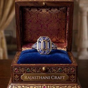 Può includere: Un anello in oro con una gemma blu centrale taglio smeraldo circondata da pietre blu rettangolari più piccole, in un cofanetto di legno con rivestimento in velluto blu e la scritta RAJASTHANI CRAFT con dettagli floreali in oro.